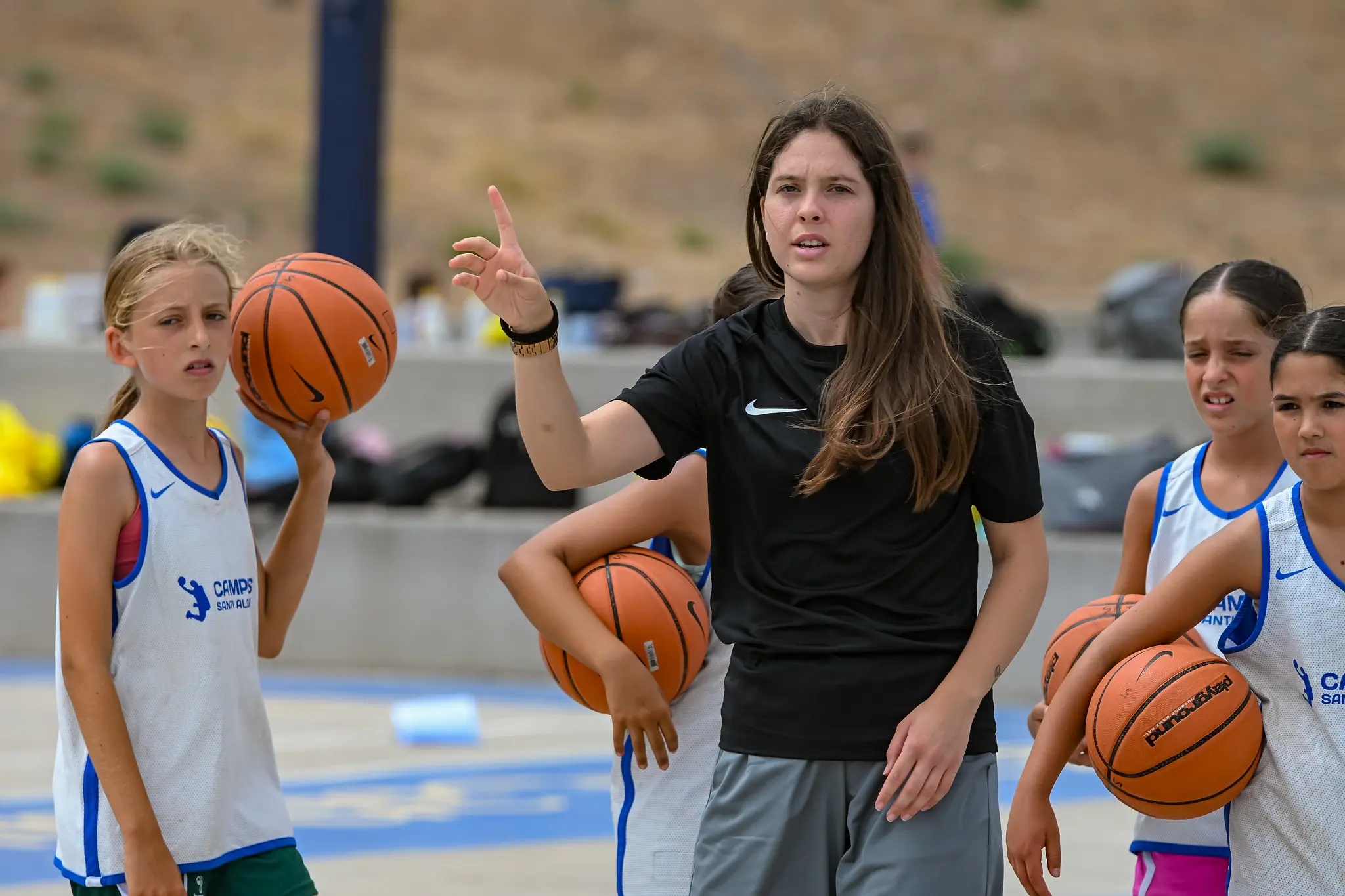 Entrenadora del Campus Santi Aldama explicando un ejercicio a un grupo de jóvenes jugadoras con balón en la pista exterior