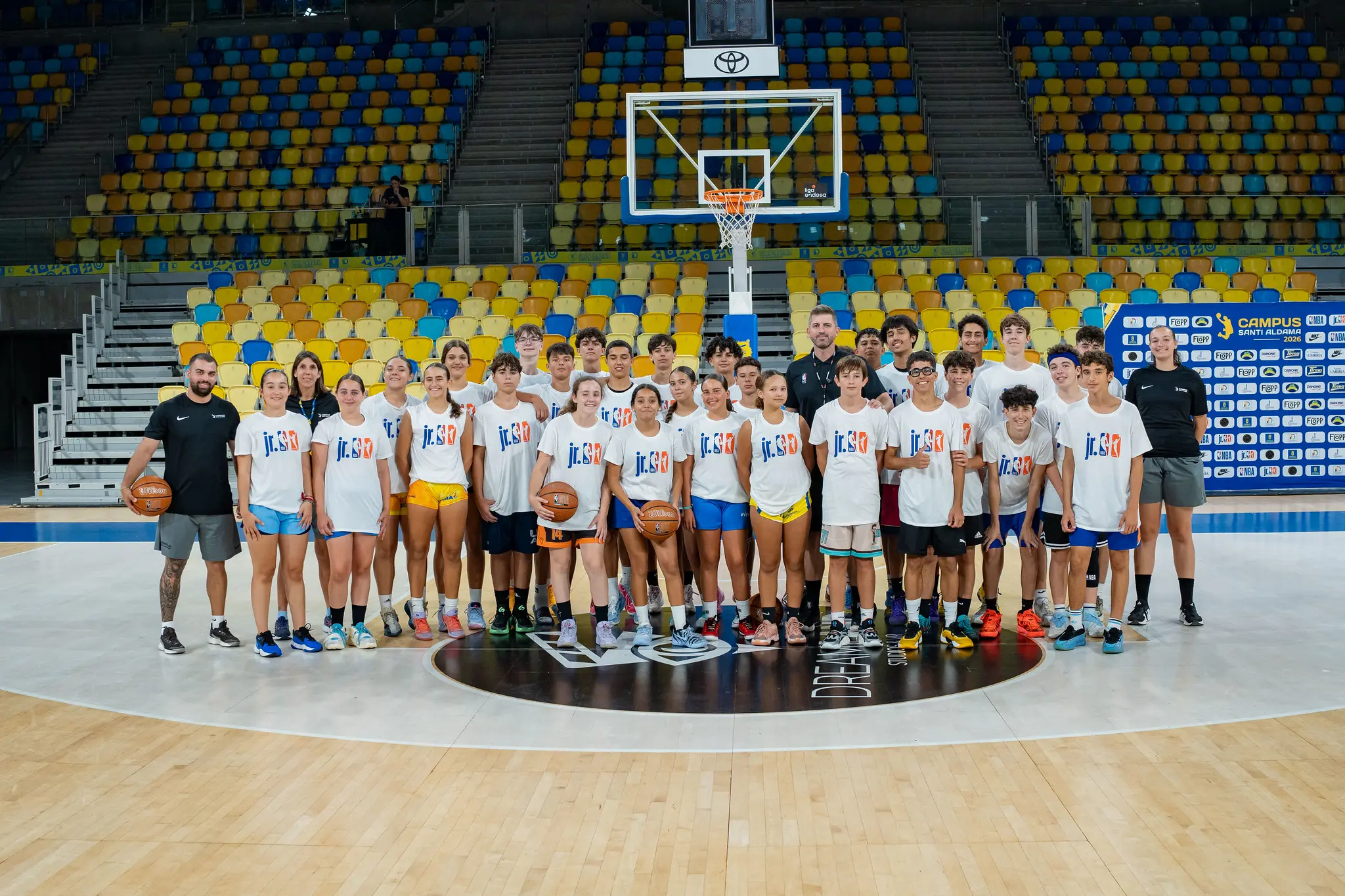 Foto de grupo de los alumnos y entrenadores del Campus Santi Aldama con camisetas Jr. NBA en el centro de la cancha del Gran Canaria Arena