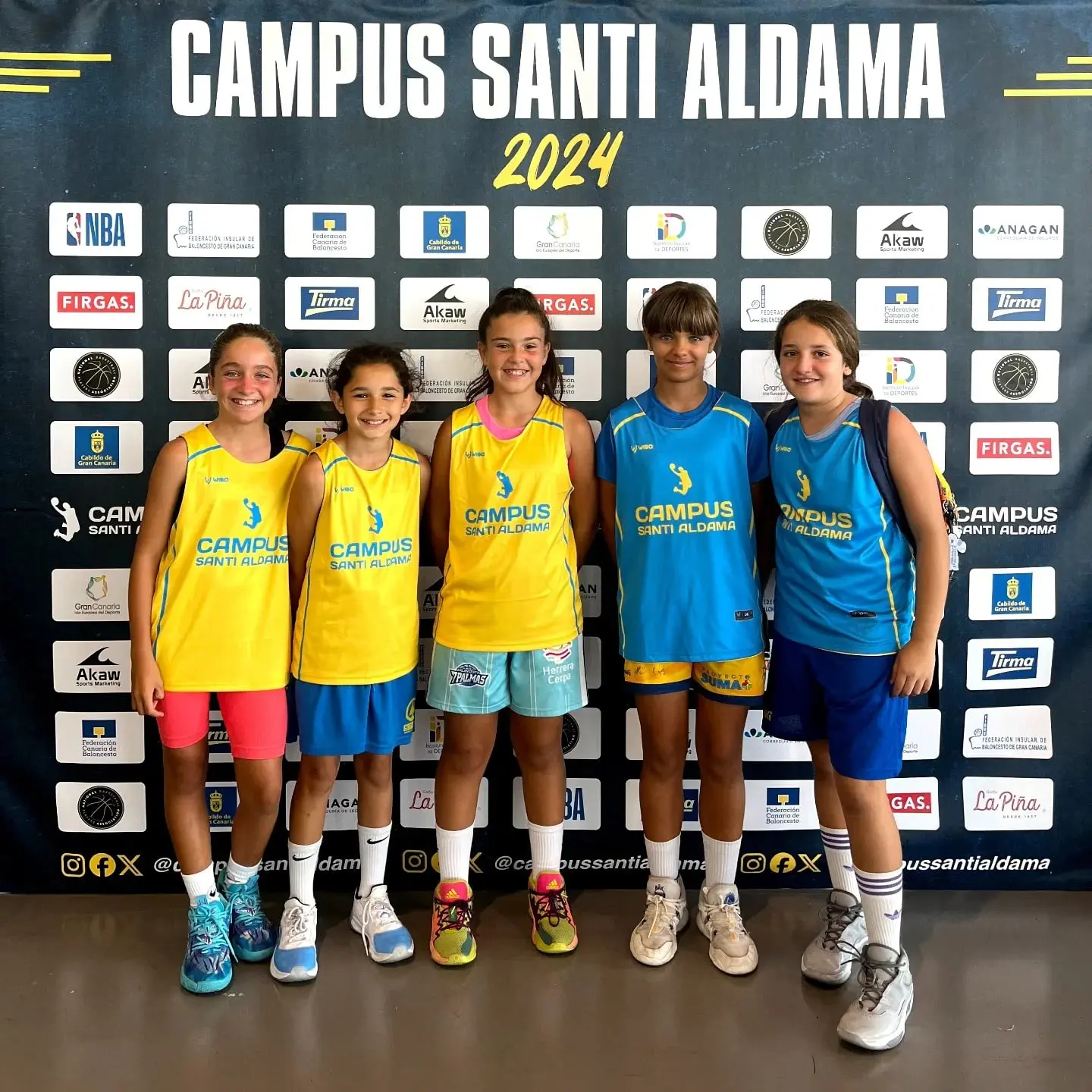 Cinco jóvenes jugadoras del Campus Santi Aldama posando sonrientes con las equipaciones oficiales frente al muro de patrocinadores